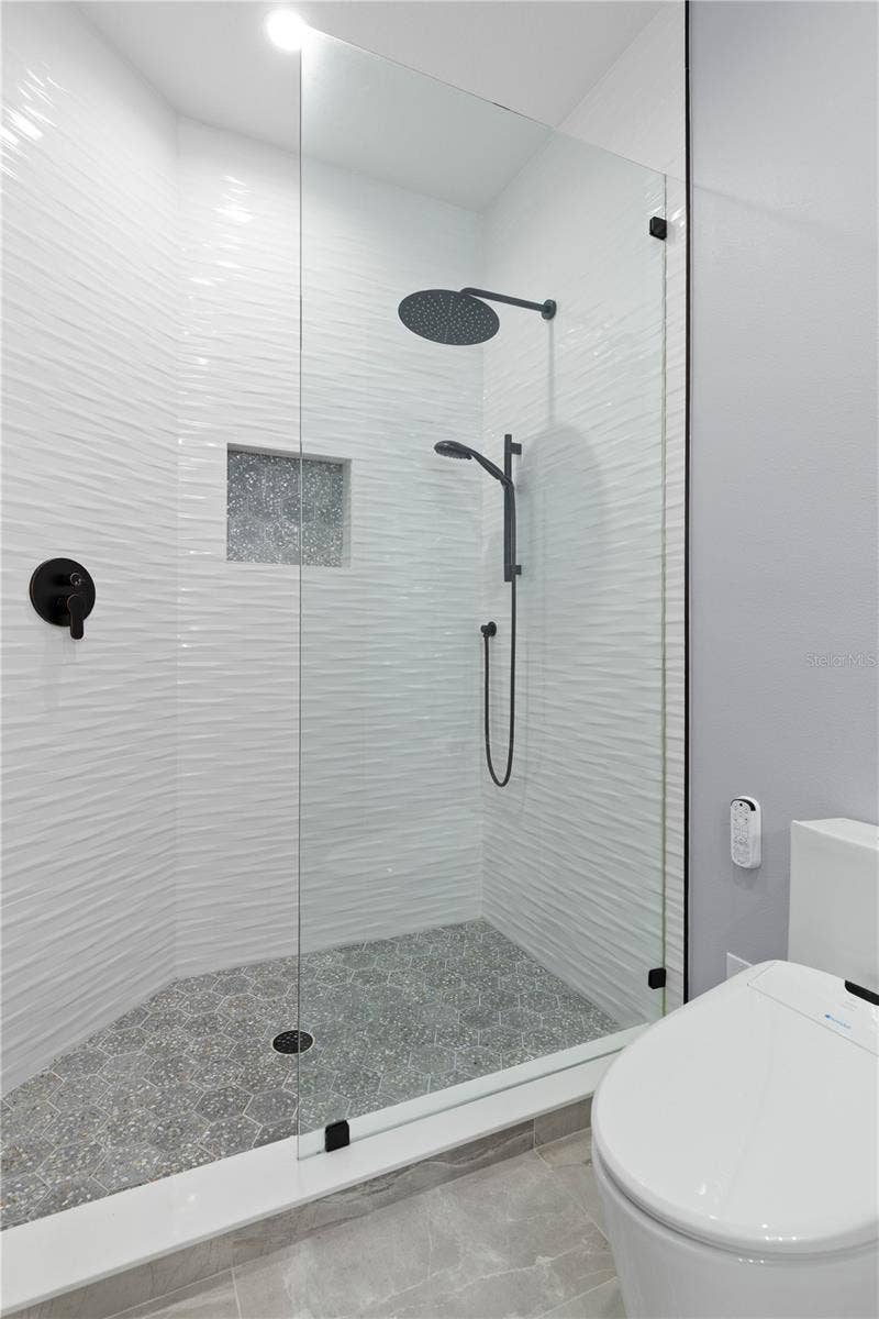The ensuite continues with a glass-enclosed walk-in shower, rainfall and handheld showerheads, and striking textured tile for a crisp, contemporary finish.