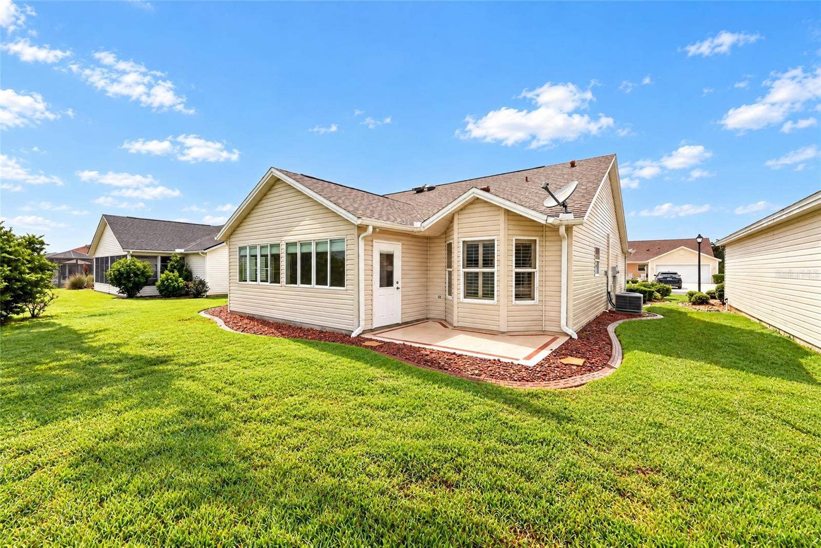 3064 BURBANK LANE, The Villages, FL 32162 Rear exterior showcasing a spacious lawn, mature landscaping, attractive rooflines, and a convenient rear entry with a decorative patio area.