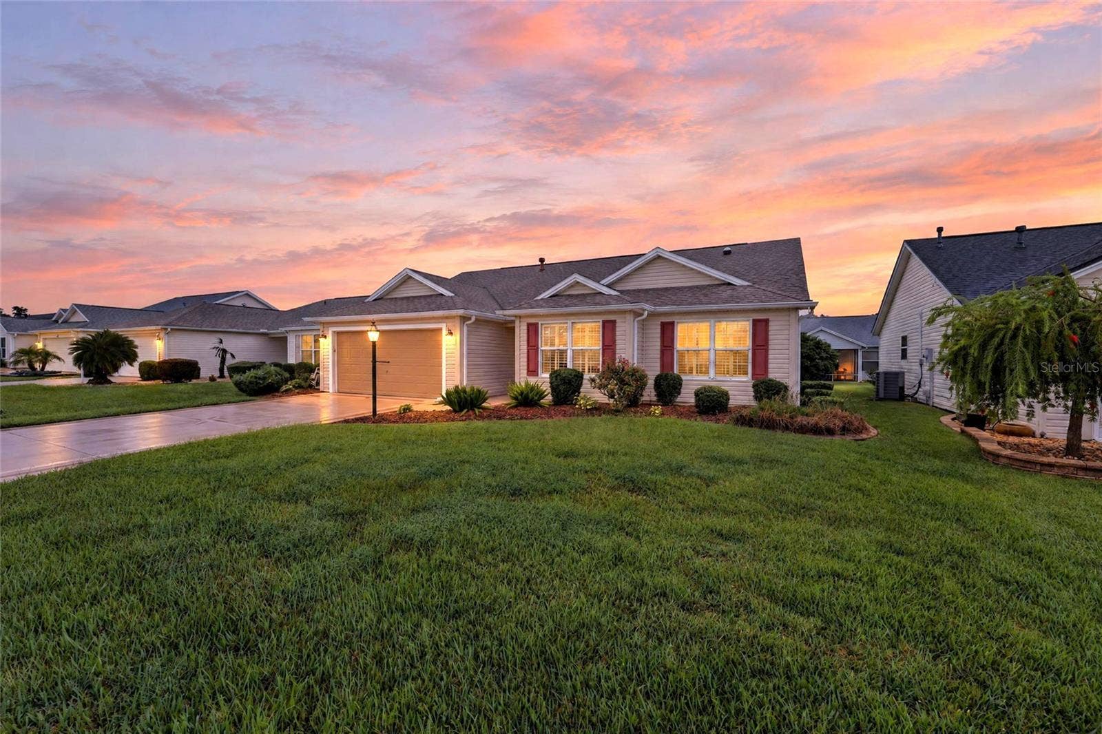 3064 BURBANK LANE, The Villages, FL 32162 Beautiful twilight view showcasing the home's inviting exterior, illuminated windows, manicured landscaping, and spacious front lawn beneath a colorful sunset sky.