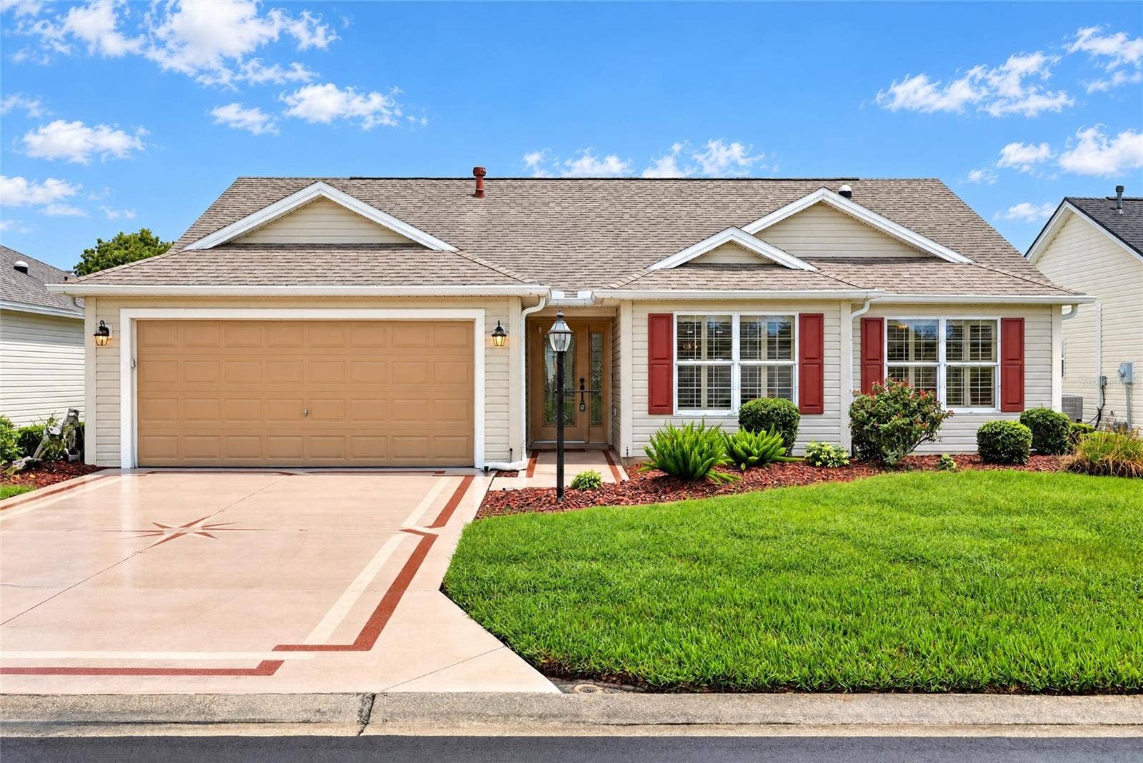 3064 BURBANK LANE, The Villages, FL 32162 Welcoming front elevation featuring a two-car garage, decorative driveway accents, attractive landscaping, and charming red shutters.
