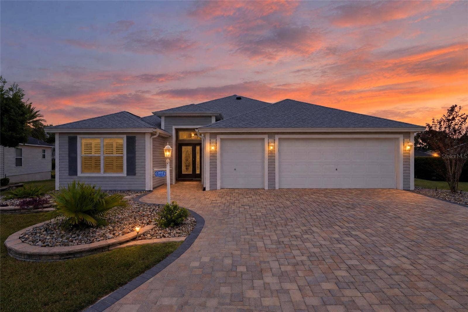 2454 MORNING DOVE LANE, The Villages, FL 32162 A glowing Florida sunset showcases this homes beautiful landscaping, sweeping paver driveway, and welcoming curb appeal.