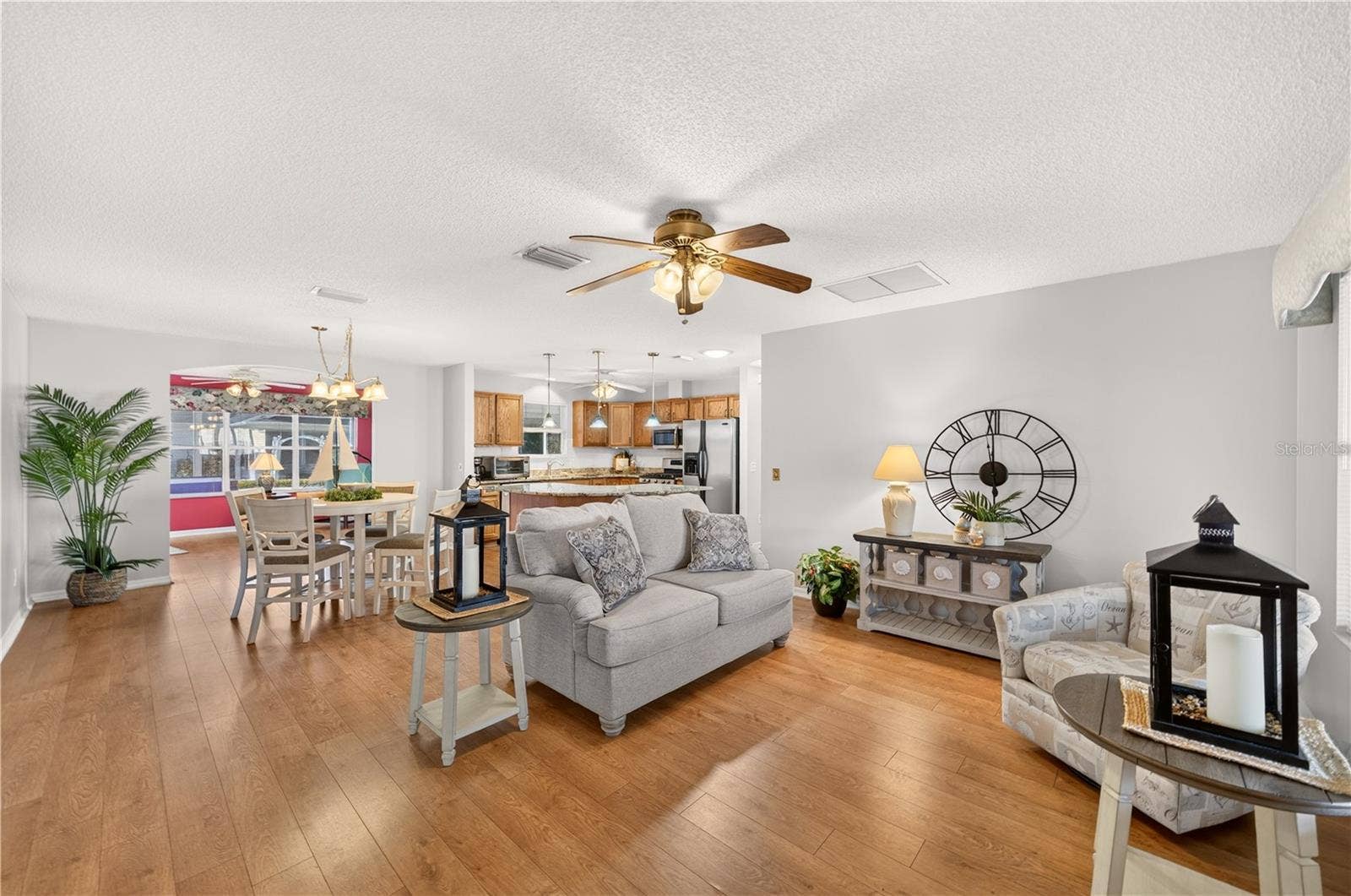 Bright and welcoming living room featuring attractive wood-look flooring, neutral wall colors, a ceiling fan, and an open layout connecting the dining area and kitchen.