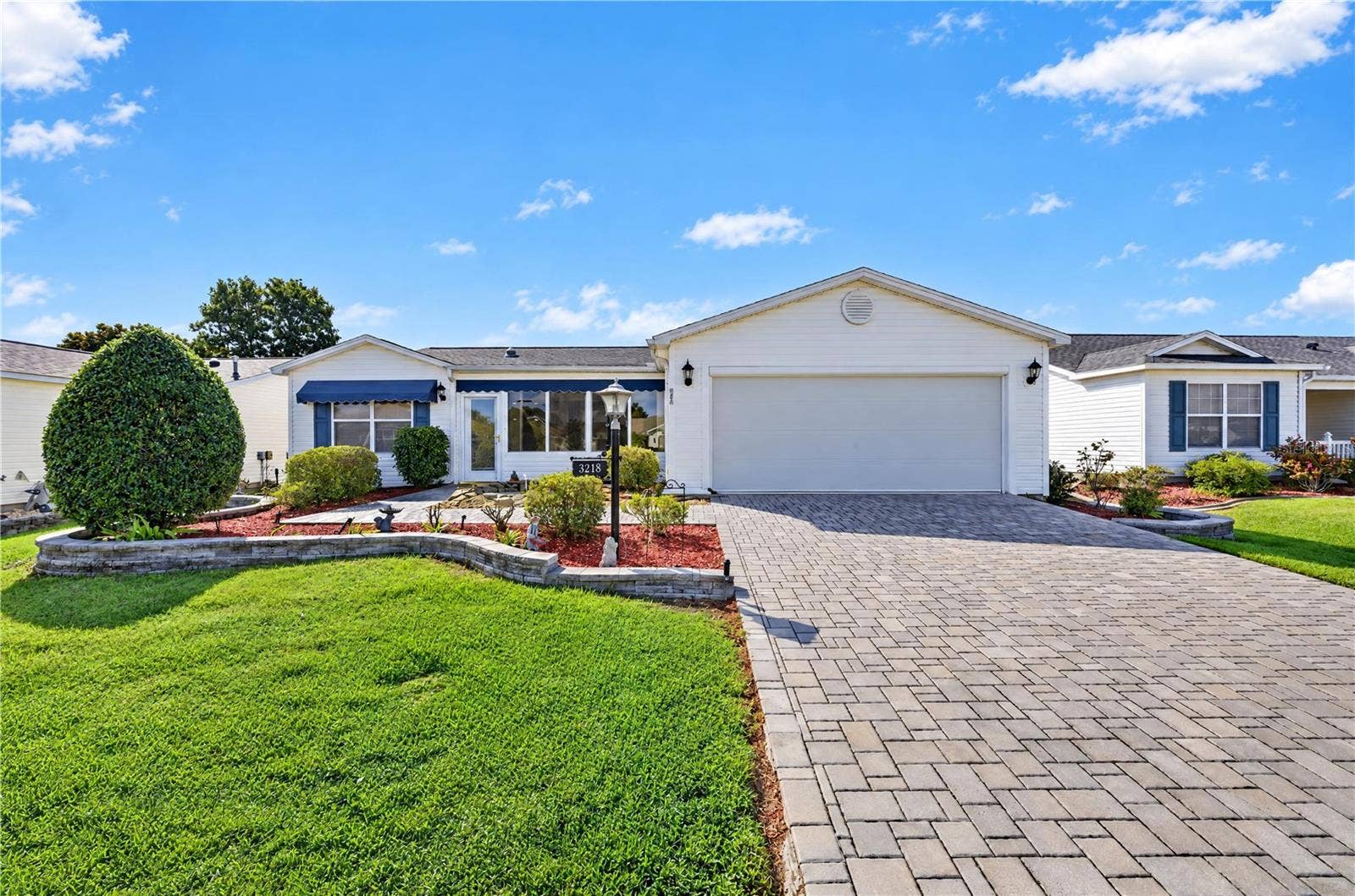 Inviting front exterior featuring a spacious two-car garage, expansive brick-paver driveway, manicured landscaping, decorative stone borders, and distinctive blue awnings.