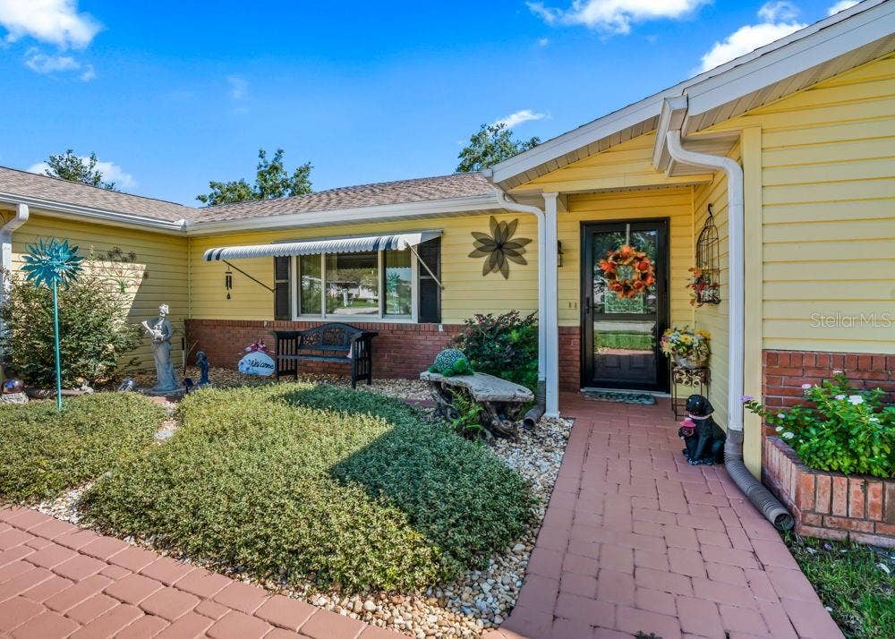 17909 SE 99TH AVENUE, Summerfield, FL 34491 Welcoming covered entry with a brick-paver walkway, cheerful yellow siding, and classic brick accents. Landscaped garden beds, a cozy bench beneath a striped window awning, and decorative touches create a charming first impression.