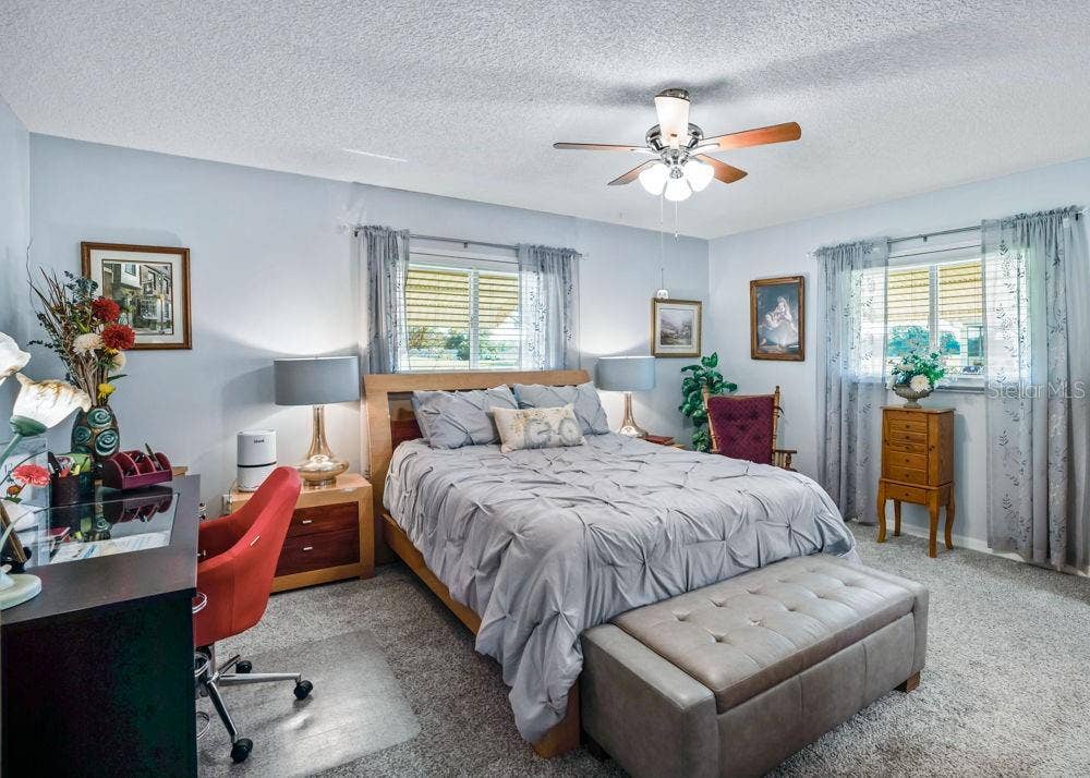 17909 SE 99TH AVENUE, Summerfield, FL 34491 Spacious primary bedroom featuring soft blue walls, carpeting, and a ceiling fan with lighting. Two windows welcome natural light, while the generous layout offers room for a desk or cozy reading area.