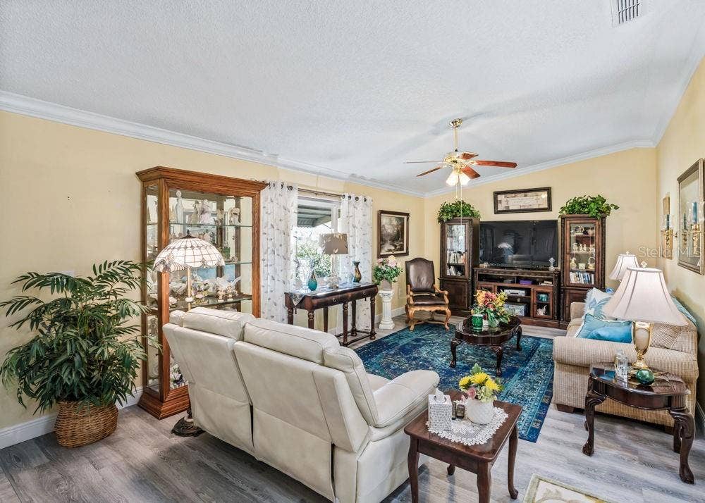 17909 SE 99TH AVENUE, Summerfield, FL 34491 Bright and inviting living room featuring wood-look vinyl flooring, crown molding, and a ceiling fan. A large window welcomes natural light, while soft neutral walls create a comfortable setting for relaxing or entertaining.