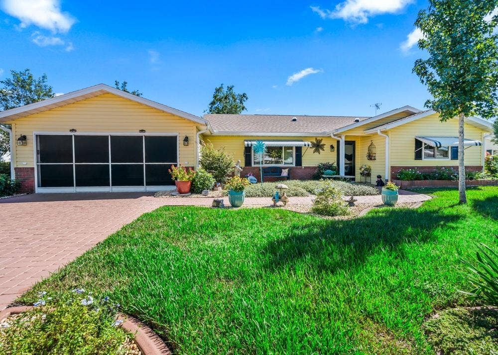 17909 SE 99TH AVENUE, Summerfield, FL 34491 Charming front exterior with cheerful yellow siding, brick accents, a screened garage, and a brick-paver driveway. A covered front porch, striped window awnings, and colorful landscaped beds add inviting curb appeal.