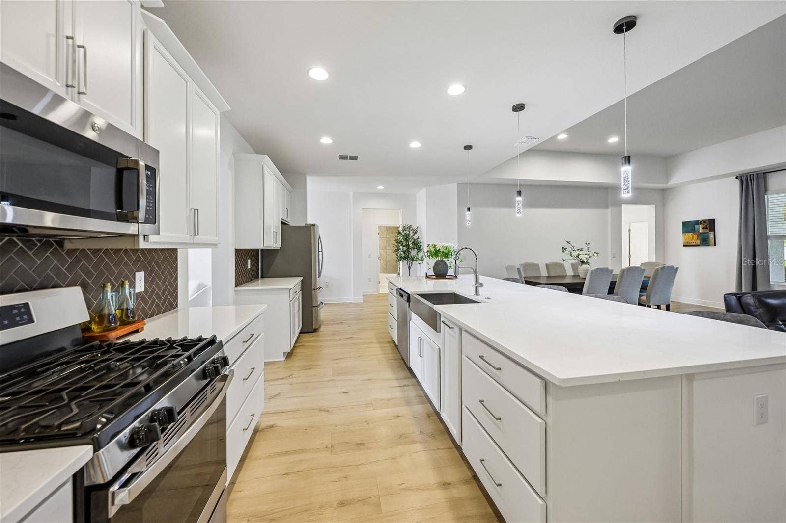 A massive QUARTZ ISLAND with bar seating sits beneath designer PENDANT LIGHTING, while STAINLESS STEEL APPLIANCES  including a GAS RANGE  pair perfectly with the stylish herringbone tile backsplash.