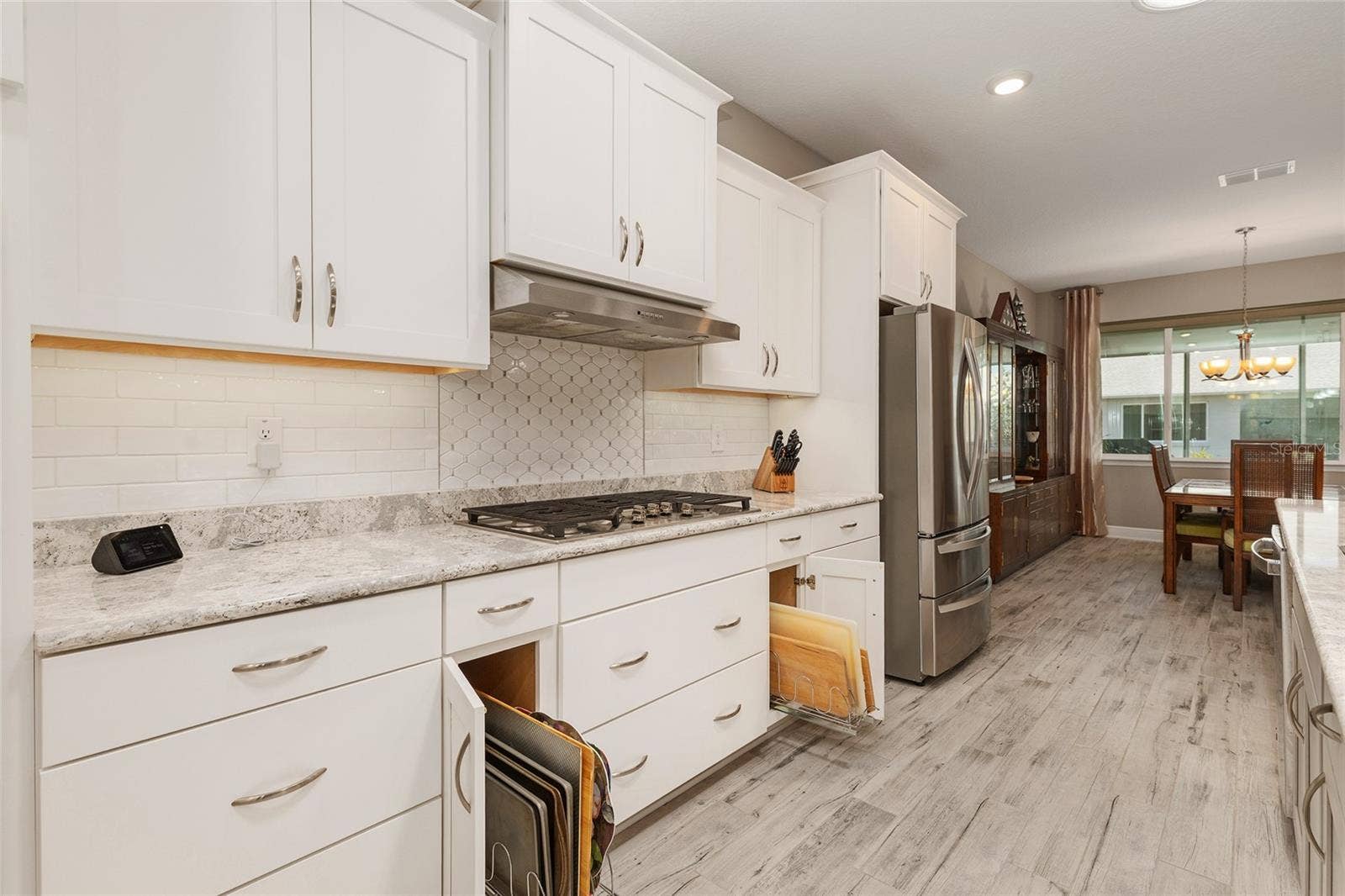 Thoughtfully designed kitchen featuring convenient pull-out storage organizers for cutting boards and baking sheets, Cambria quartz countertops, decorative tile backsplash, over- and under-cabinet lighting, stainless steel appliances, and an open flow into the dining area.