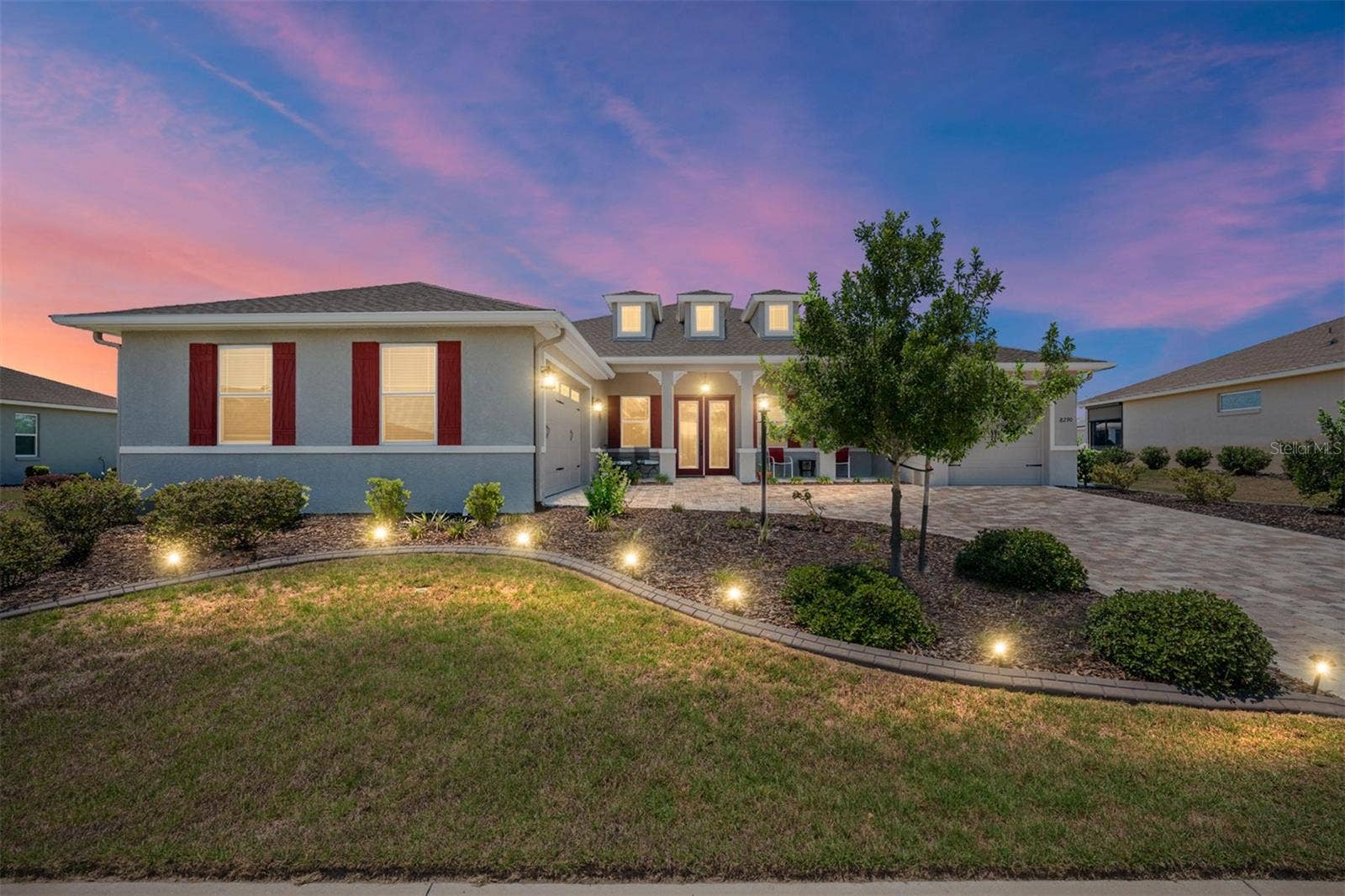 Stunning twilight view highlighting the home's exceptional curb appeal, beautifully illuminated landscaping, expansive paver driveway, and welcoming covered front porch. The elegant leaded-glass double entry doors and warm exterior lighting create an inviting first impression.