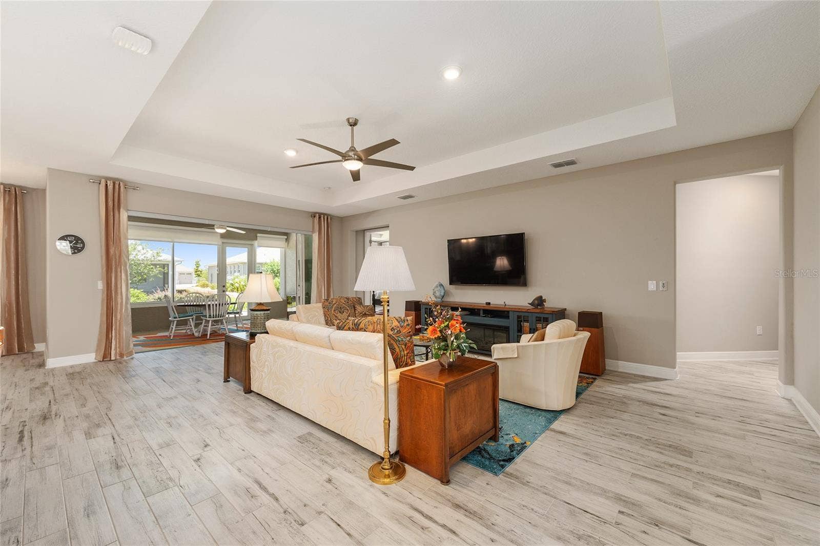 Expansive great room featuring a tray ceiling, recessed lighting, ceiling fan with integrated lighting, ceramic plank flooring, and stacking sliding glass doors with a zero-threshold transition to the enclosed lanai. An inviting space for relaxing and entertaining, with the wall-mounted TV included in the sale.