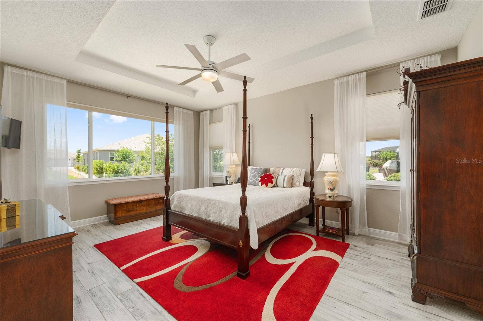 Spacious first primary suite, currently used as the owner's suite, featuring an elegant tray ceiling, ceiling fan with integrated lighting, ceramic plank flooring, neutral finishes, and multiple windows that fill the room with natural light. Wall-mounted TV included in the sale.