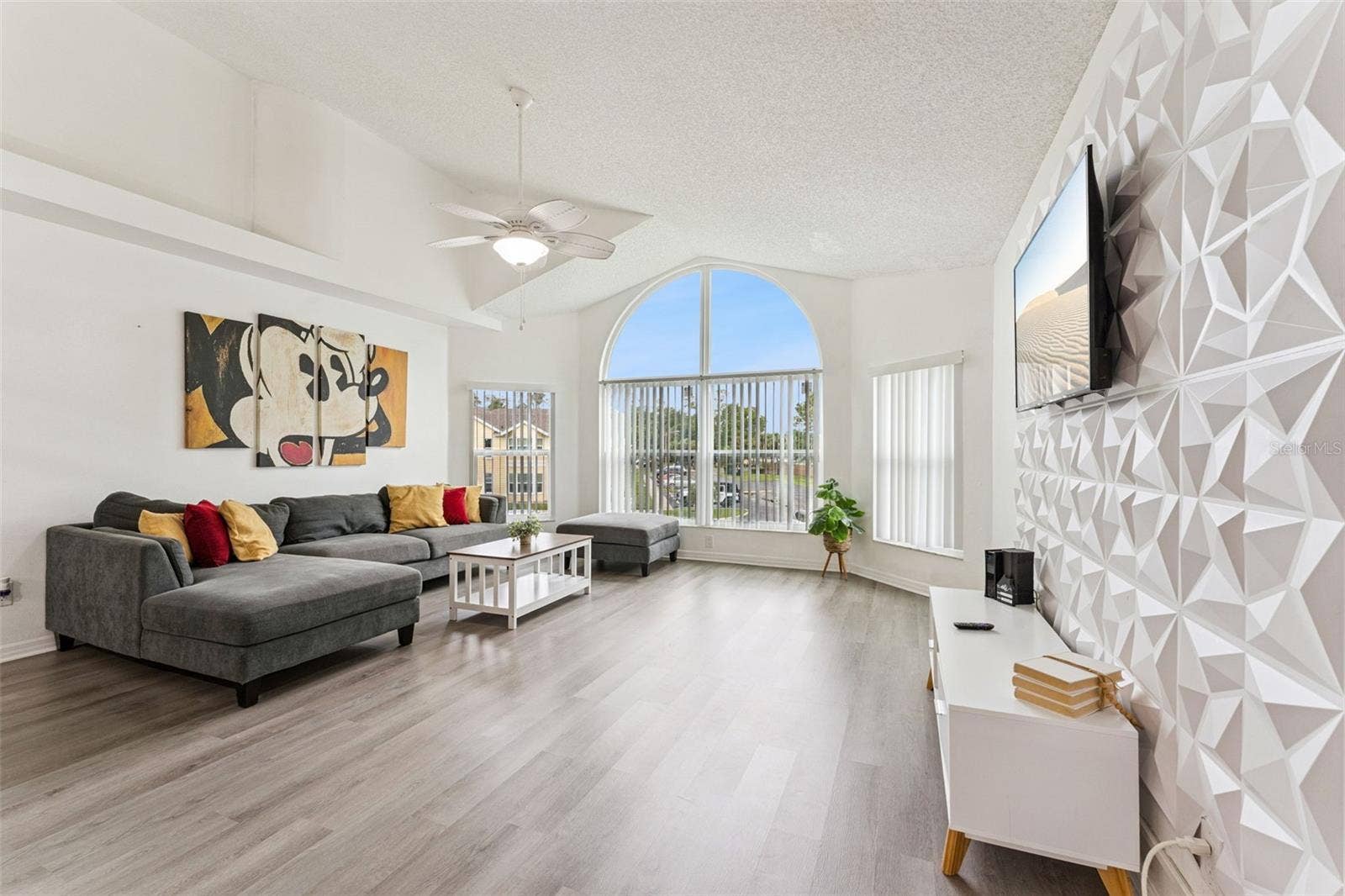 5017 LAGUNA BAY CIRCLE, #100, Kissimmee, FL 34746 Living Room: Sun-drenched living room boasting vaulted ceilings, a geometric 3D feature wall, wood-look plank flooring, and a large architectural arched window