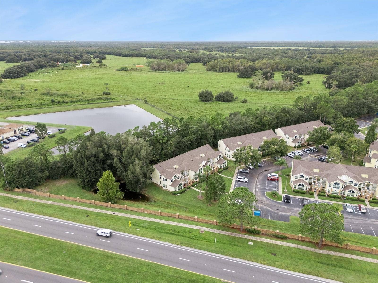 5017 LAGUNA BAY CIRCLE, #100, Kissimmee, FL 34746 Water Feature & Open Fields Aerial: Wide aerial perspective featuring the community's adjacent tranquil pond, wooded buffer line, and rolling green backdrop.
