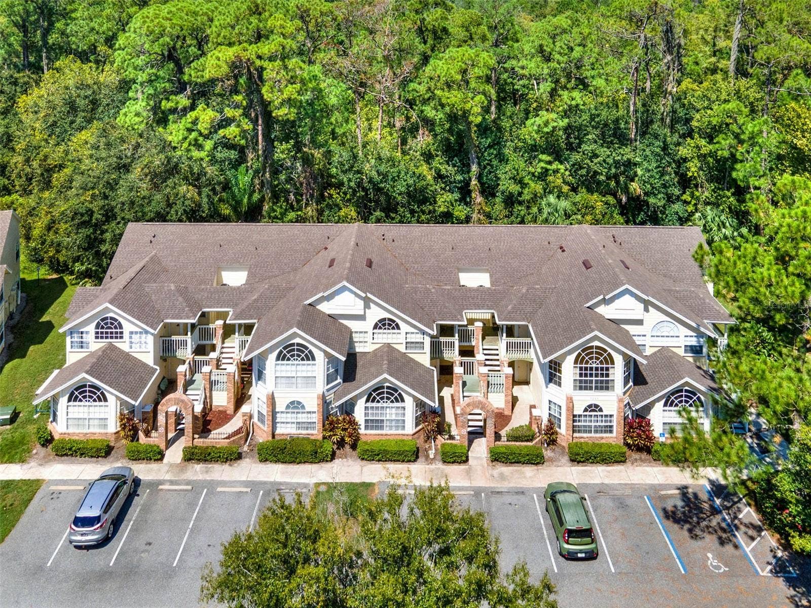 5017 LAGUNA BAY CIRCLE, #100, Kissimmee, FL 34746 uilding Exterior Aerial: Direct front aerial perspective showcasing the unit building, parking spaces, brick entry archways, and dense wooded tree canopy behind.