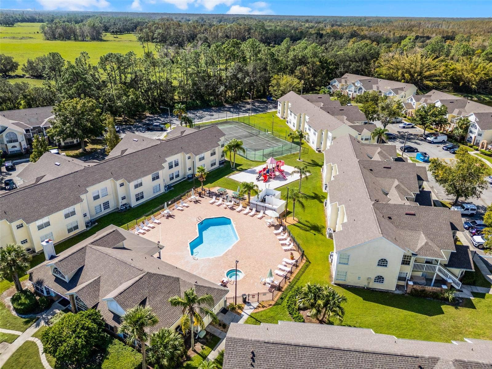5017 LAGUNA BAY CIRCLE, #100, Kissimmee, FL 34746 Amenity Hub Aerial: Elevated aerial view highlighting the central pool deck, clubhouse, sand playground, tennis court, and surrounding residential buildings set against open green spaces.