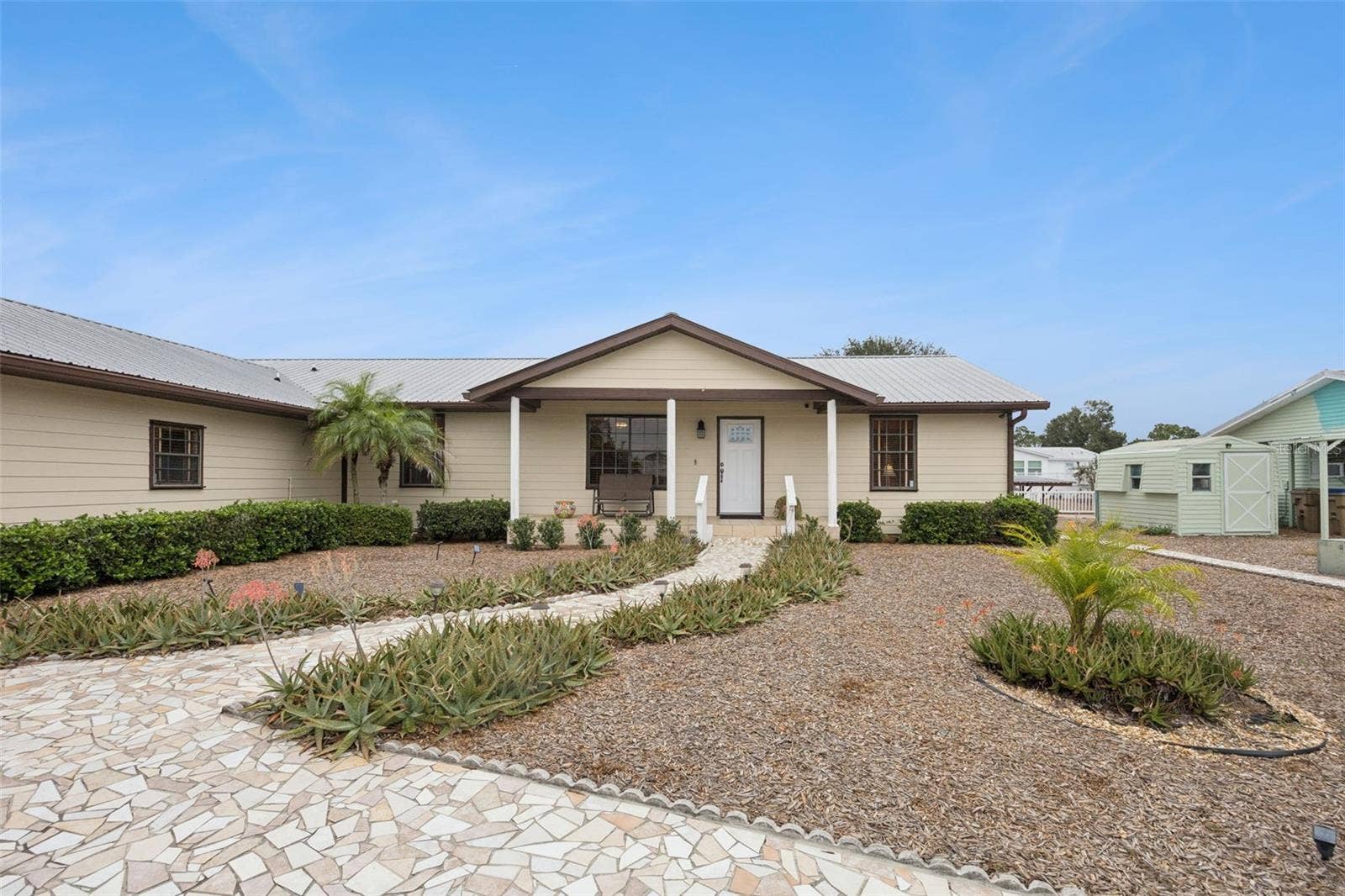 Exterior Front Elevation: Welcoming ranch-style front exterior featuring stone-paved walkways, low-maintenance gravel landscaping, a durable metal roof, and a covered entry porch.