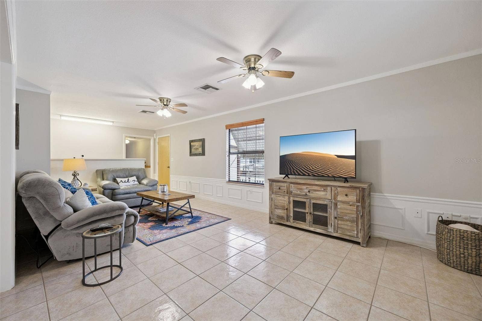 Main Living Room: Expansive open living room featuring neutral ceramic tile flooring, classic wainscoting trim, ceiling fans, and built-in media area.