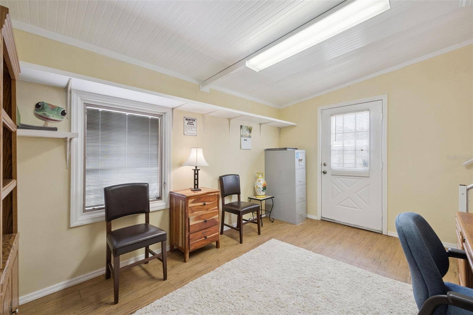 Home Office / Flex Space: Functional office or hobby room featuring wood-look vinyl flooring, overhead strip lighting, built-in shelving, and exterior door access.