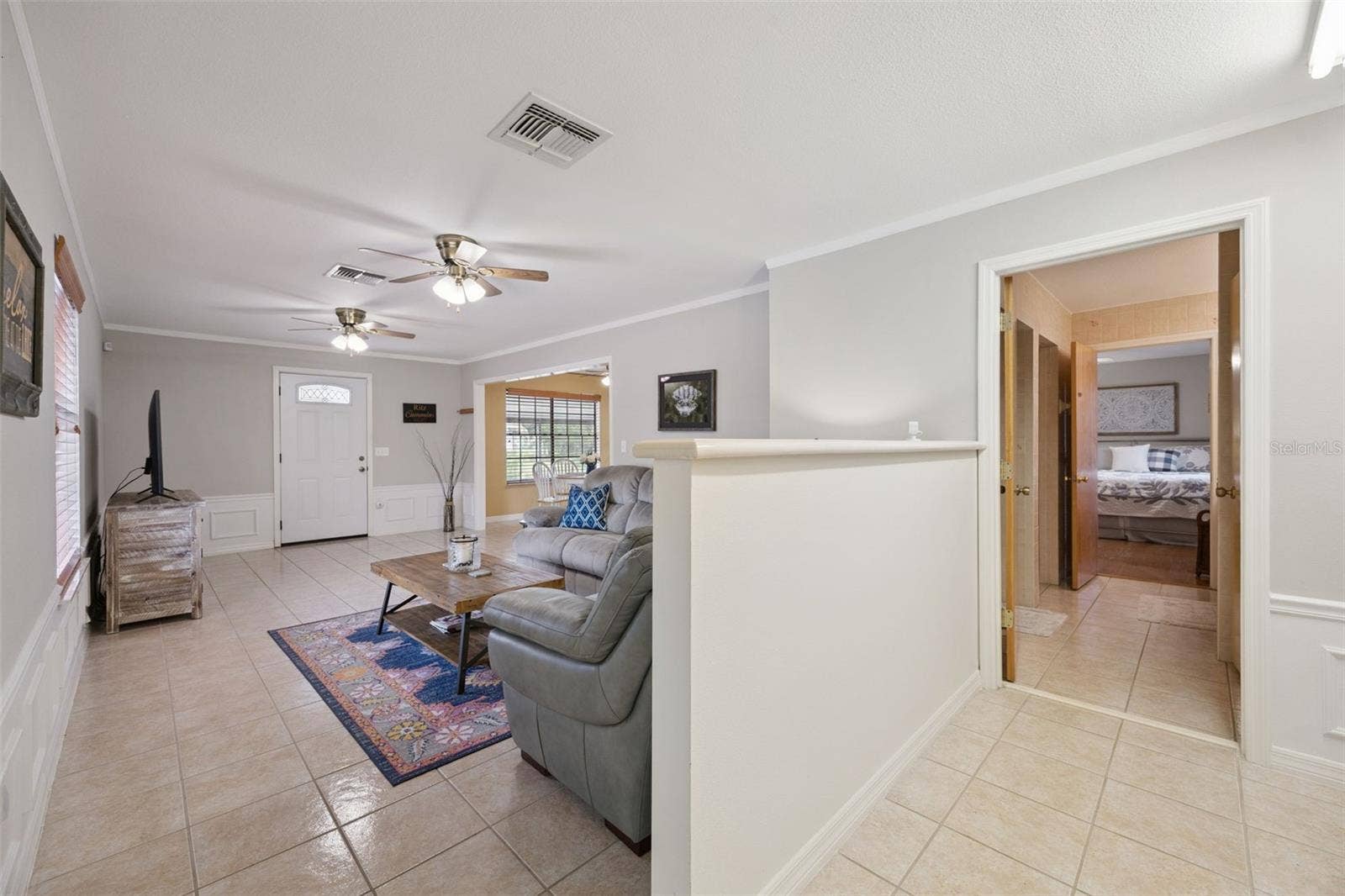Living Room & Hall Transition: Broad living space detail showing tile floors, half-wall divider, wainscoting trim, and doorway access toward secondary rooms.