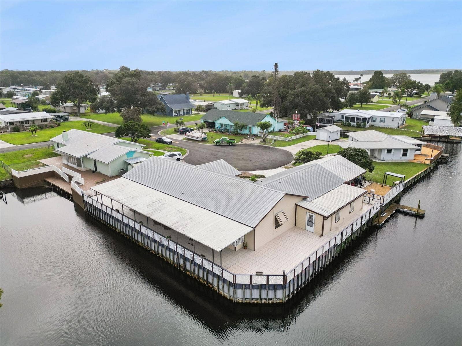 Aerial Waterfront Corner View: High-angle drone shot capturing the full perimeter of the home's wraparound waterfront deck and dock area.