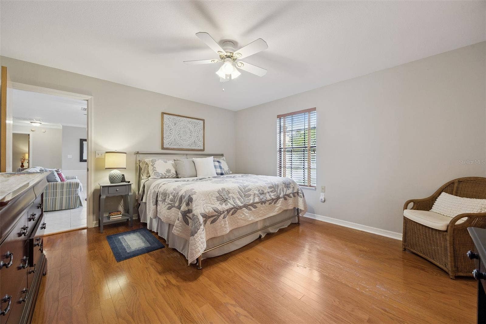 Primary Bedroom Layout: Generous primary suite showing wood-look flooring, large window with blinds, ceiling fan, and seamless connection to main living areas.