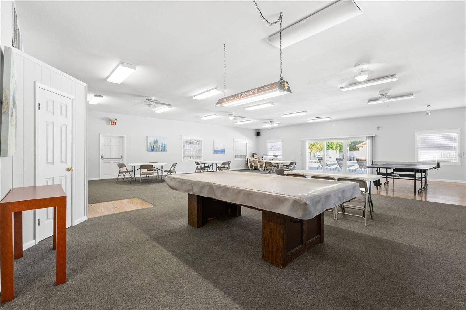 12760 INDIAN ROCKS ROAD, #107, Largo, FL 33774 The clubhouse offers a large gathering space with billiards, ping-pong and multiple tables for games, cards or community events, with doors opening toward the pool area beyond.