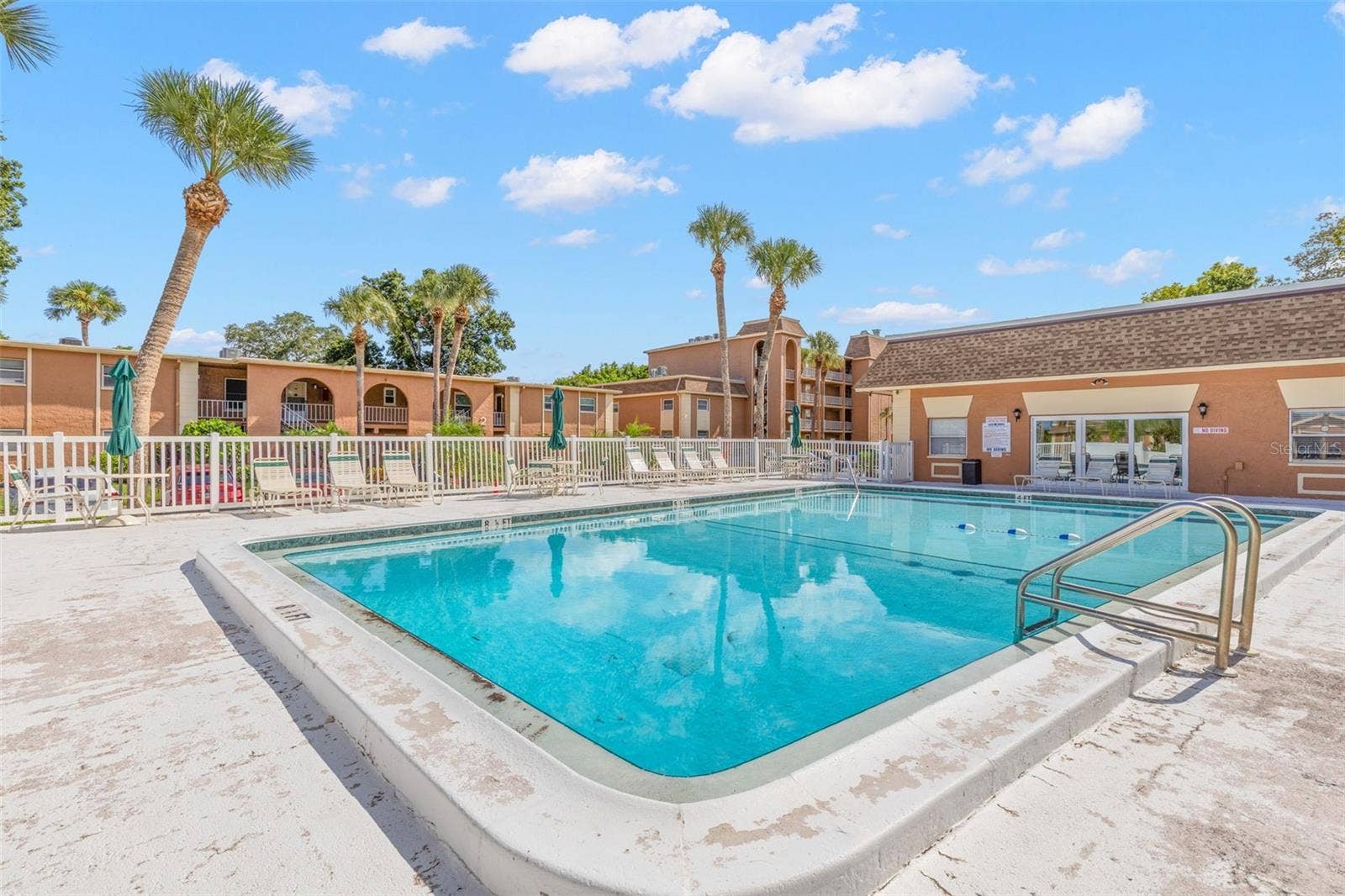 12760 INDIAN ROCKS ROAD, #107, Largo, FL 33774 The community pool is surrounded by a spacious deck with lounge seating and umbrellas, while palms and the neighboring clubhouse create an inviting spot to relax outdoors.
