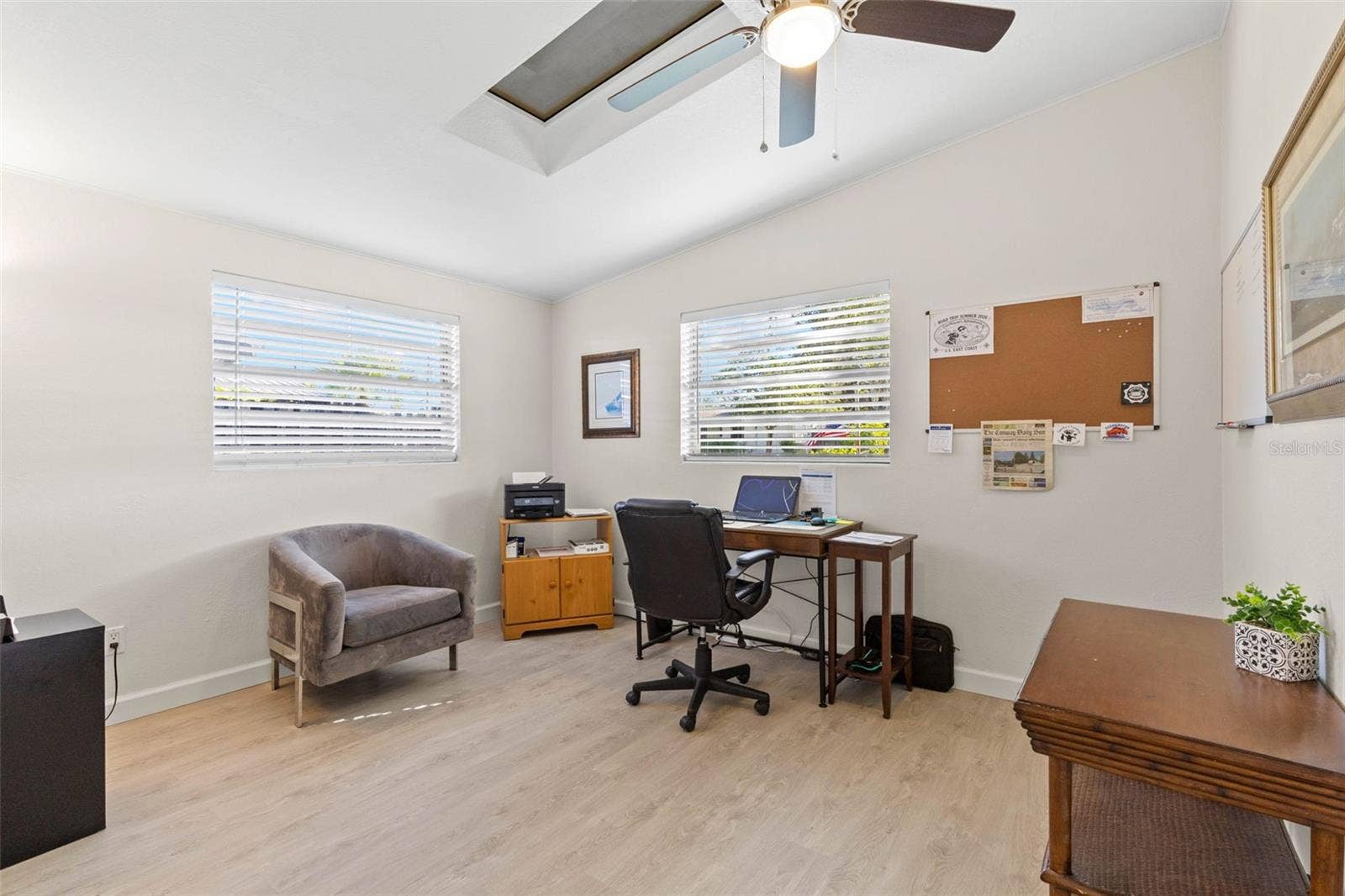 A bright secondary bedroom, currently used as a home office, offers two windows, a skylight, ceiling fan, neutral tones, and luxury vinyl plank flooring (2025), with plenty of flexibility for guests, work, or hobbies.