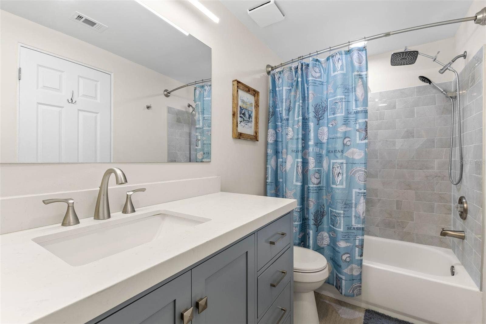 The remodeled primary bath (2025) pairs a wide gray vanity and sleek white countertop with a tiled tub/shower surround, rainfall showerhead, handheld sprayer, and updated fixtures for a clean, modern finish.
