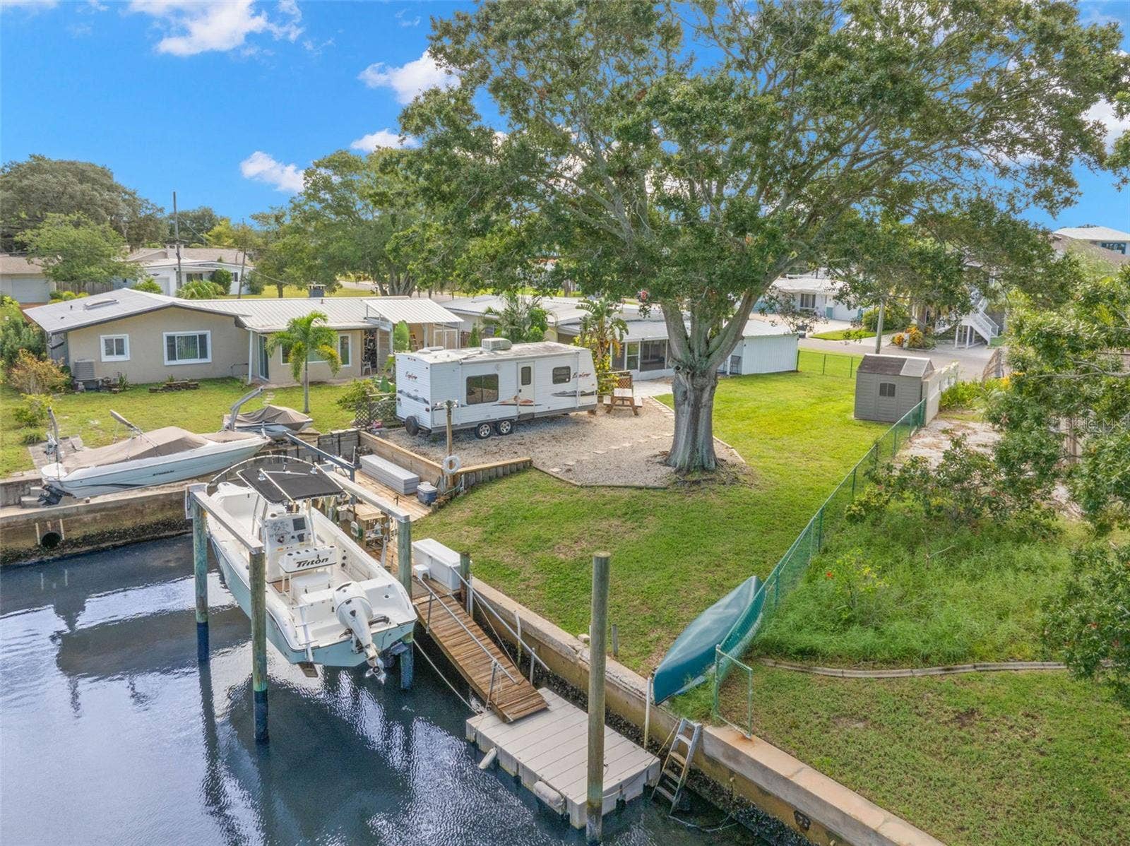An aerial view showcases the propertys waterfront lifestyle, with a 10,000-pound boat lift, wooden dock and floating dock along the canal, plus generous lawn space, RV parking, and mature shade trees just steps from the water.