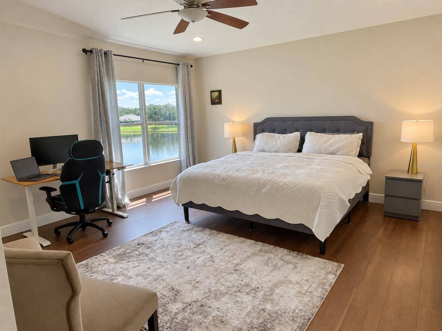 A spacious bedroom with water views, a large window, and room for both a sitting area and a desk. An upholstered bed, soft neutral finishes, and a ceiling fan complete the space.