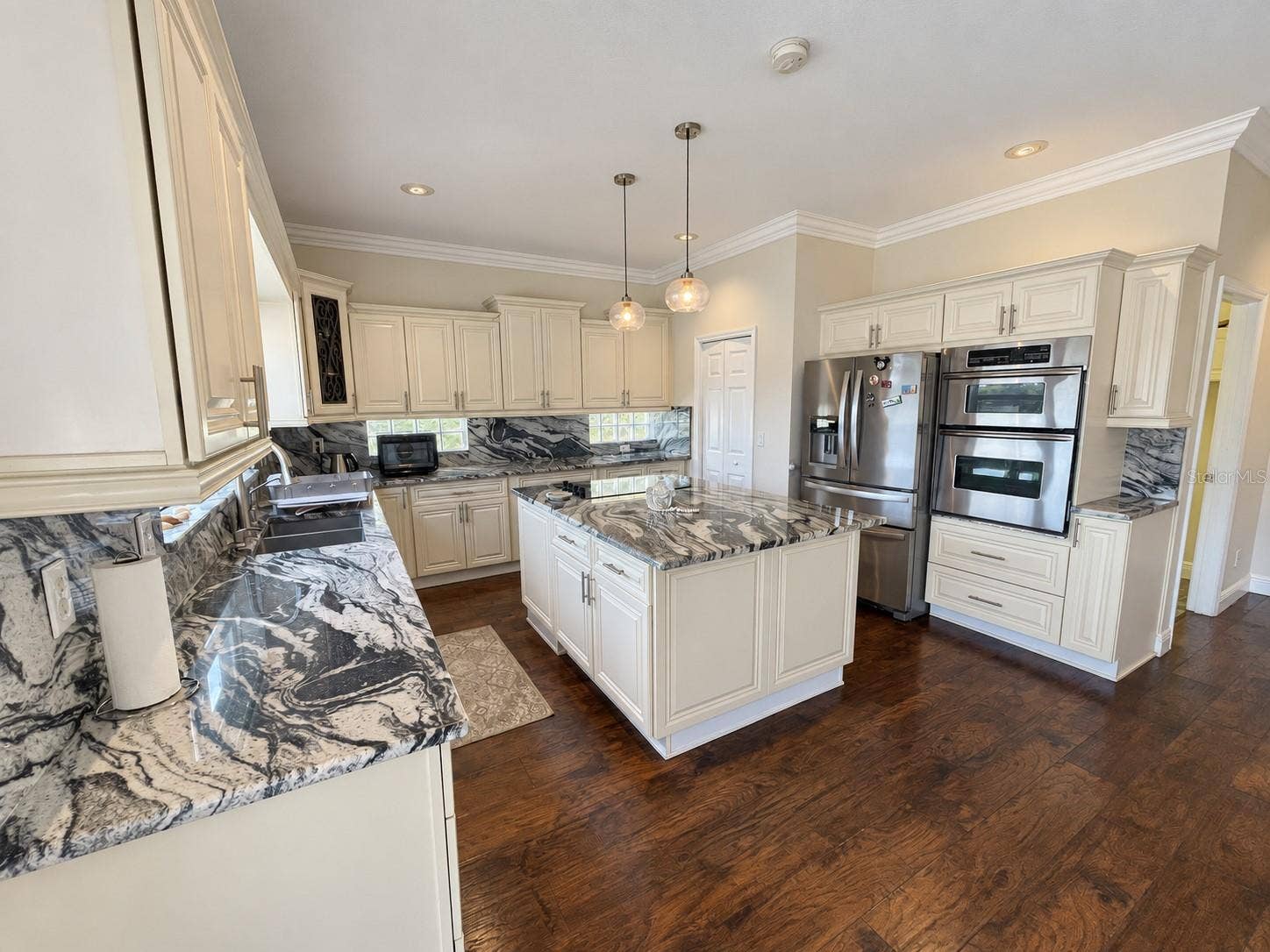 Spacious kitchen featuring cream cabinetry, striking stone countertops and matching backsplash, a central island with an electric cooktop, and stainless steel appliances, including double wall ovens. A large window above the sink overlooks the backyard and water.