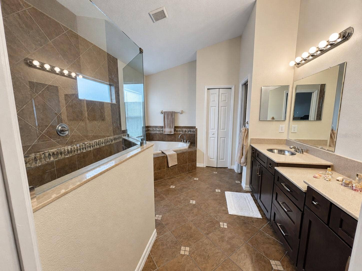 A generous bathroom featuring two sinks, extensive vanity storage, a separate soaking tub, and a walk-in shower with glass panels. High ceilings, natural light, and a separate toilet compartment enhance the layout.
