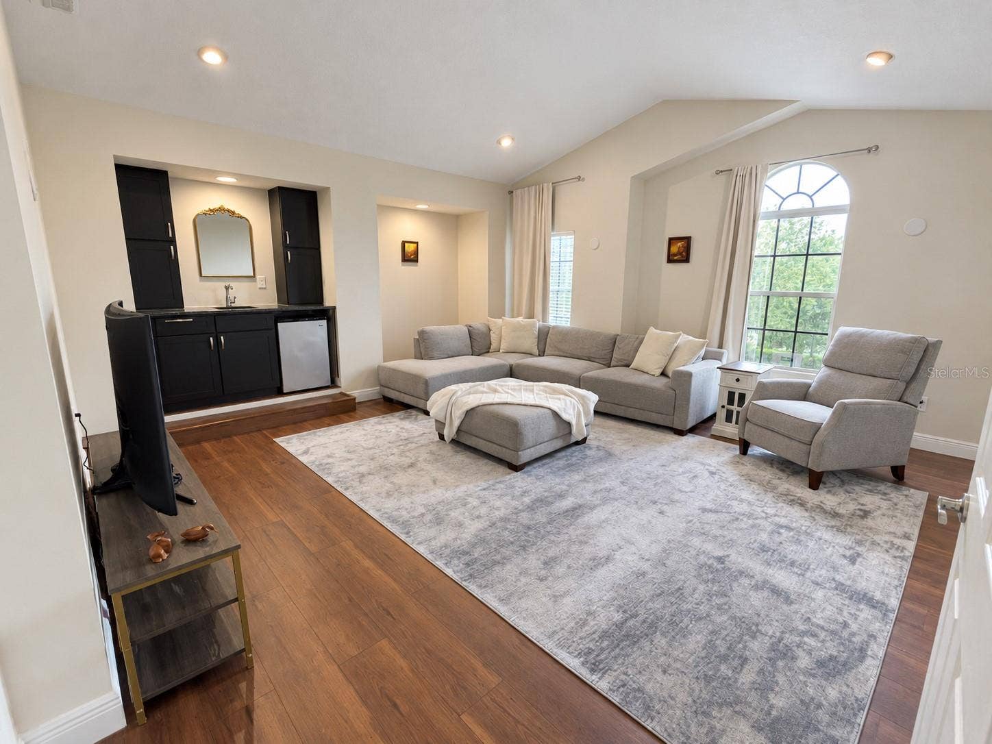 A spacious retreat with vaulted ceilings, tall windows, and room for a sectional and additional seating. A built-in wet bar with cabinetry and a compact refrigerator makes the room convenient for relaxing and entertaining.