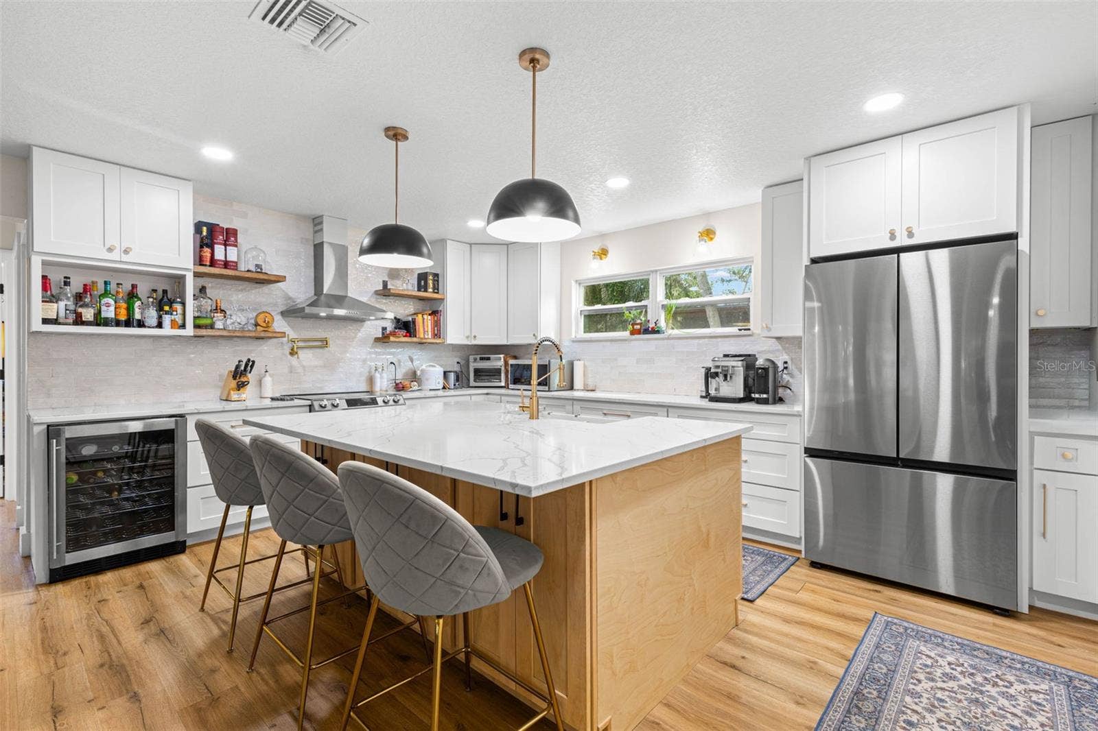 3013 GRAHAM LANE, Tampa, FL 33618 This beautifully designed kitchen combines style and practicality with abundant white shaker cabinetry, quartz countertops, stainless steel appliances, a built-in beverage refrigerator, and a generously sized center island. Floating wood shelves, gold accents, and contemporary pendant lighting add character and warmth.