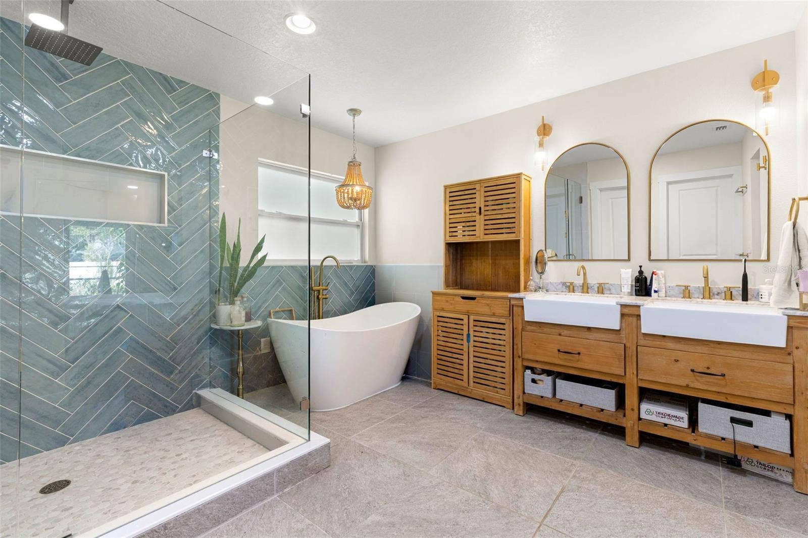 3013 GRAHAM LANE, Tampa, FL 33618 The stunning primary ensuite offers a spa-inspired retreat featuring a freestanding soaking tub, an oversized walk-in shower with frameless glass enclosure, dramatic blue-green herringbone tile, and dual farmhouse-style vessel sinks set atop warm wood vanities. Gold fixtures, arched mirrors, and coordinating sconces complete this beautifully designed bathroom.