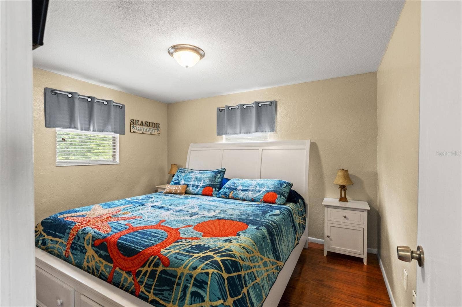 A comfortable bedroom with warm wood flooring, neutral walls, and two windows providing natural light. The room accommodates a generously sized bed with nightstands, while its coastal-inspired dcor complements the home's relaxed waterfront setting.