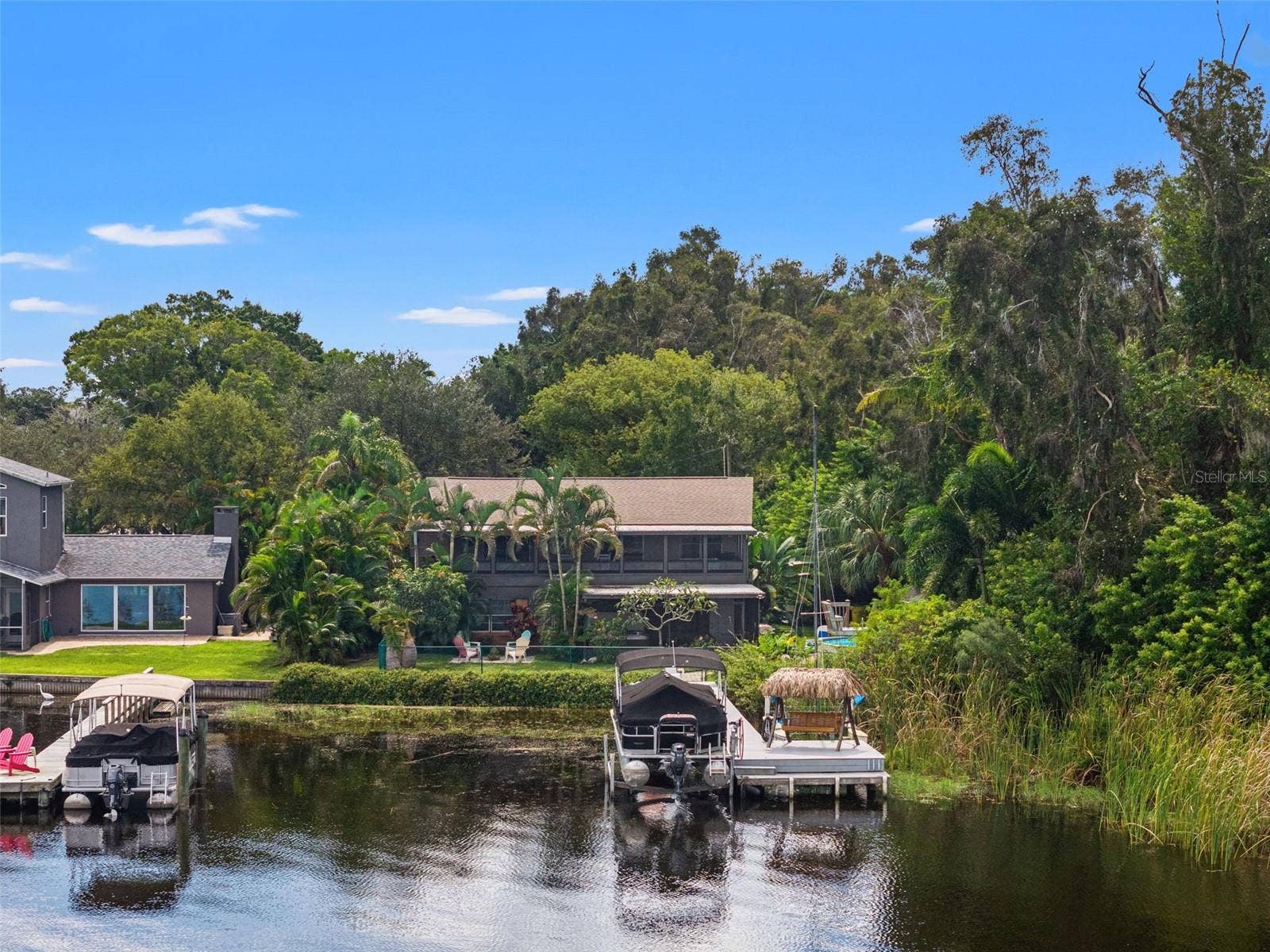 The home's waterfront setting is particularly appealing from this perspective. A private dock with a pontoon boat lift and thatched-roof swing sits along the shoreline, while dense vegetation borders one side of the property, creating a sense of separation from neighboring homes.
