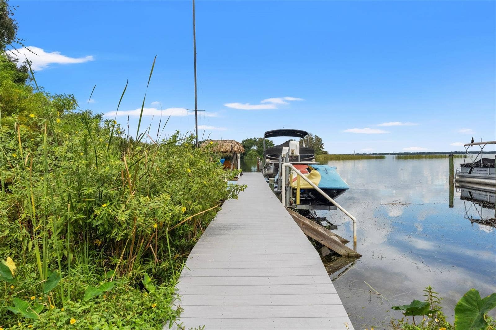 A walkway leads directly to the waterfront dock, where a boat and personal watercraft are pictured alongside the calm lake. With expansive water views stretching toward the distant shoreline, this is an inviting setting for boating, fishing, and enjoying life on the water.