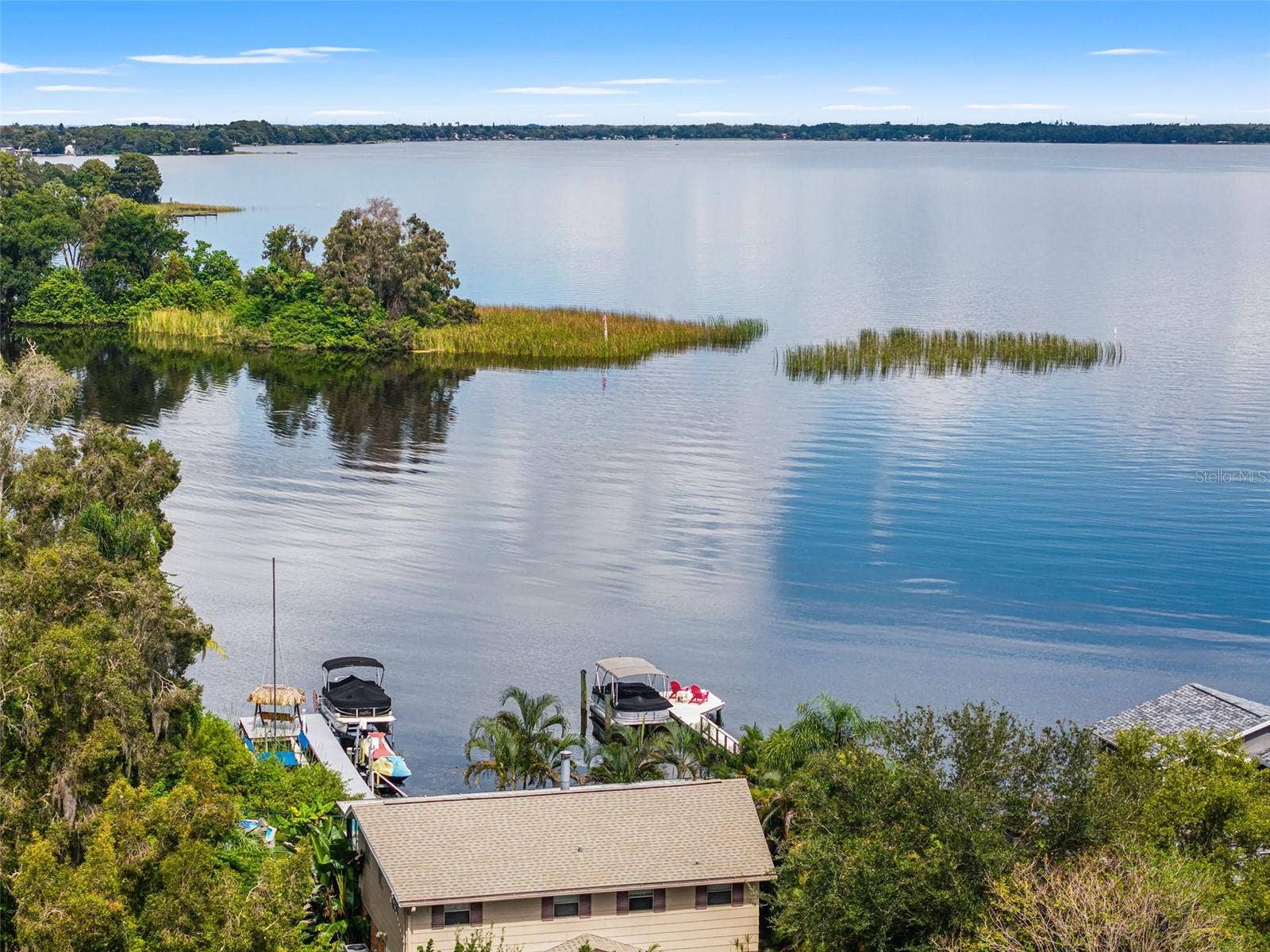 An elevated perspective highlights the property's dock, pontoon boat lift, and thatched-roof seating area. Beyond the shoreline, expansive open water stretches toward the distant tree line, offering an inviting setting for boating, fishing, and enjoying the outdoors.