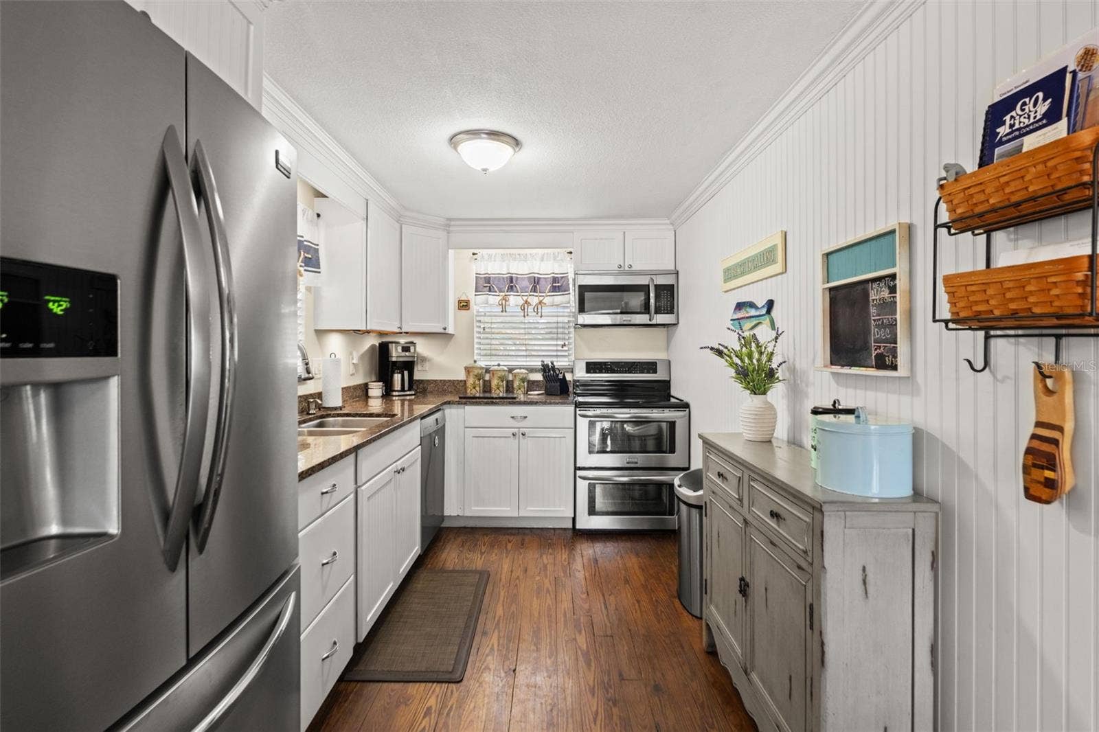 This inviting kitchen combines white cabinetry, stainless steel appliances, wood flooring, and warm-toned countertops. Crown molding and beadboard-style wall detailing add character, while the galley layout keeps everything conveniently within reach.