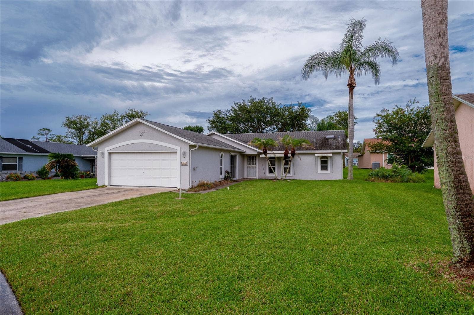 Front perspective of the home displaying a spacious front yard, two-car garage, concrete driveway, and palm trees.