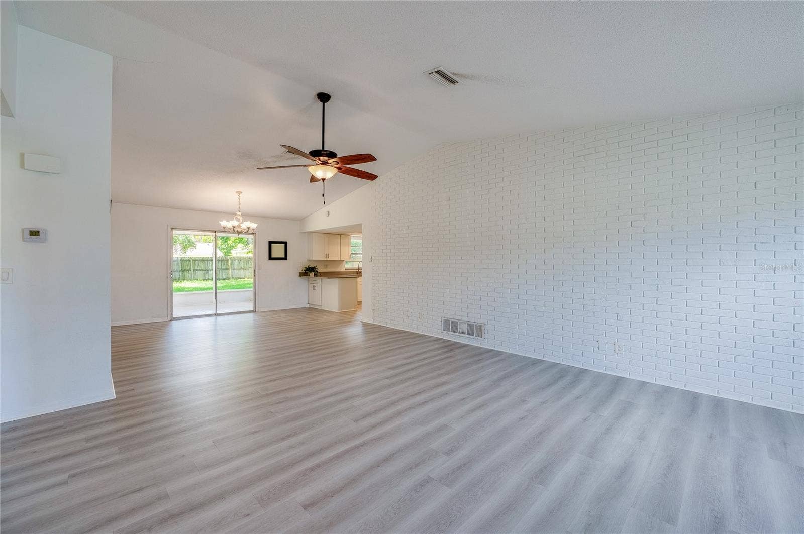 156 SPRINGWOOD DRIVE, Daytona Beach, FL 32119 Spacious open-concept living area highlighting light luxury vinyl flooring, a painted white brick accent wall, and a ceiling fan. Sliding glass doors at the rear provide seamless access and views to the backyard patio.