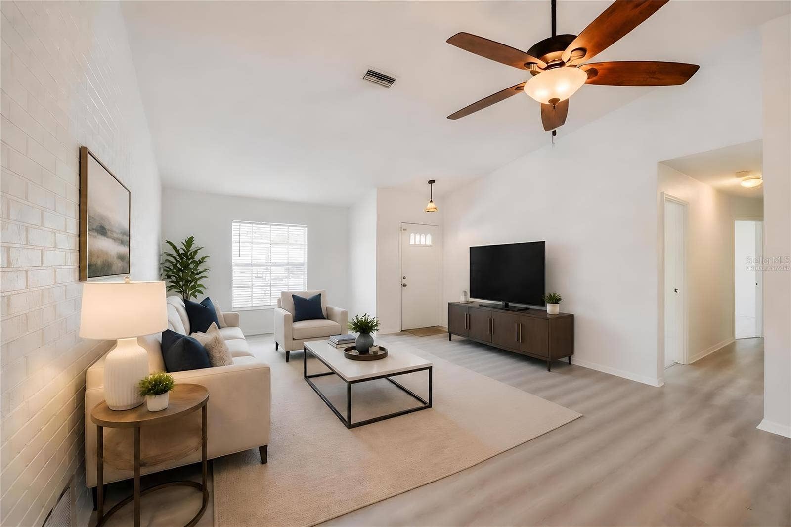 156 SPRINGWOOD DRIVE, Daytona Beach, FL 32119 Virtually Staged. Welcoming foyer and main living room showcasing white walls, modern luxury vinyl flooring, and a ceiling fan with light fixture. A painted brick accent wall adds character to the functional entryway.