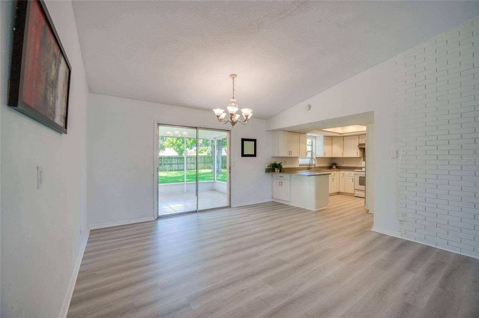 156 SPRINGWOOD DRIVE, Daytona Beach, FL 32119 Open dining and living space defined by a classic chandelier, light luxury vinyl flooring, and a white brick wall accent. Sliding glass doors open to a screened porch, filling the space with natural light.