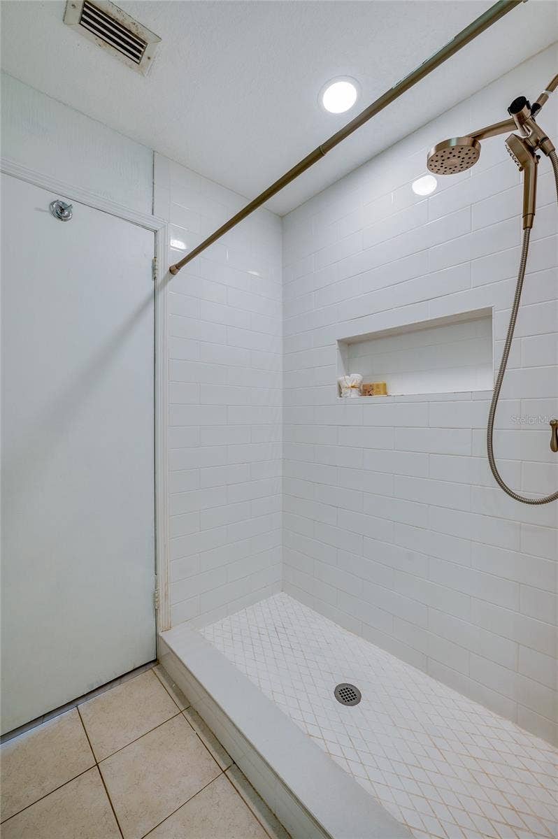 156 SPRINGWOOD DRIVE, Daytona Beach, FL 32119 Walk-in shower complete with crisp white subway tiled walls, a built-in recessed storage niche, and a bronze hand-held showerhead set over a textured tile floor basin.