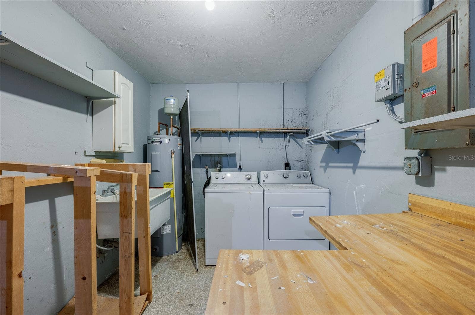 156 SPRINGWOOD DRIVE, Daytona Beach, FL 32119 Functional utility room equipped with a full-size washer and dryer, a utility sink, a water heater, and built-in shelving. Plywood work surfaces and an electrical panel offer practical workspace and storage options.