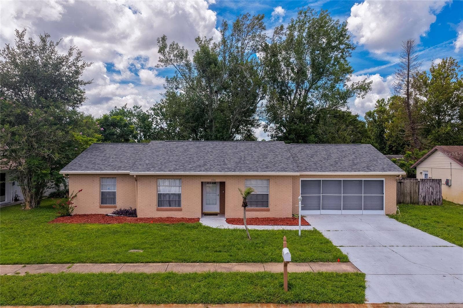156 SPRINGWOOD DRIVE, Daytona Beach, FL 32119 Charming single-story home featuring a spacious front lawn, concrete driveway, and screened garage enclosure. Lush surrounding trees and vibrant landscaping complete the inviting curb appeal.