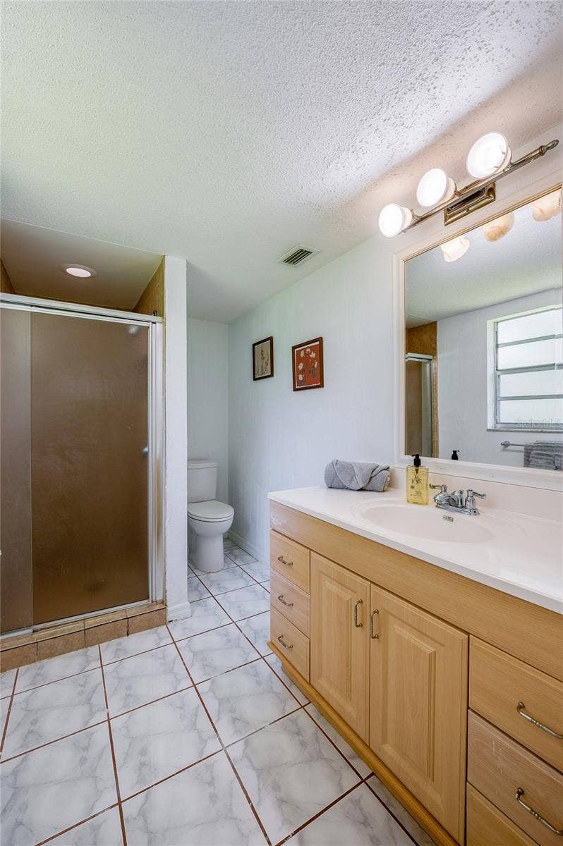 156 SPRINGWOOD DRIVE, Daytona Beach, FL 32119 En-suite bathroom featuring a light wood vanity with a long countertop, modern vanity lighting, and tiled floors. A walk-in glass shower stall and toilet area offer convenient everyday functionality.